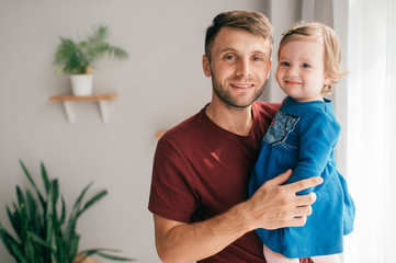 Young handsome strong dad in a bright room hold his cute little daughter which sits on a shelf