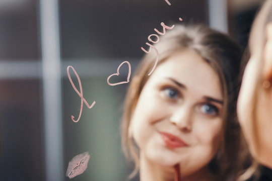 reflection of young woman face in mirror with inscription "I love you", painted heart and lip kiss, happy girl in romantic relationship, concept creative declaration of love