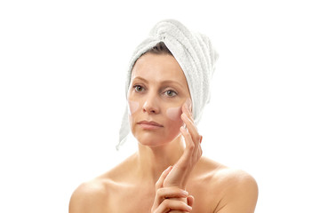 A beautiful woman with a white towel on her head does a facial rejuvenation with eye patches. White isolated background.