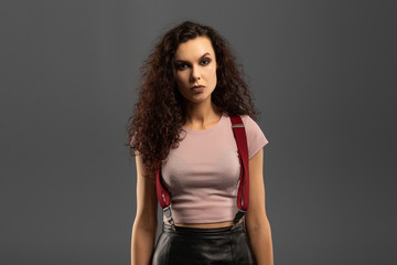 Young beautiful girl with curls on her head with a serious facial expression on a gray background. Portrait of a woman with a strong face with brown eyes in a pink T-shirt and red suspenders.