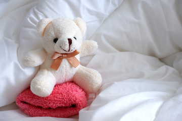 A soft toy bear, white in color, sits on a pink pillow.  White linen.