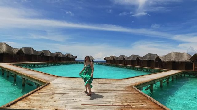 Maldive island resort. Girl in green dress walking on wooden bridge near bungalow. View of a young woman from the back. Maldives travel destination. Summer vacation in paradise. Blue ocean. Unusual
