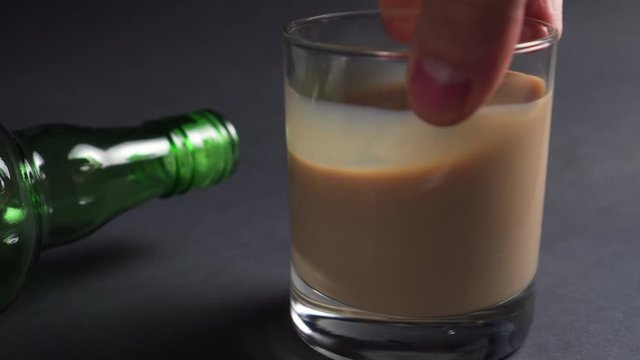 hand shakes a glass with irish cream liquor and ice cubes on a dark background with an empty alcohol bottle. close-up