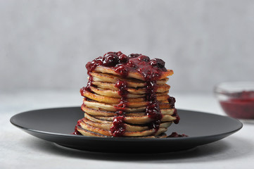 Pancakes with berry jam on a black plate. Close-up. Light background. In the background is a cup of jam.