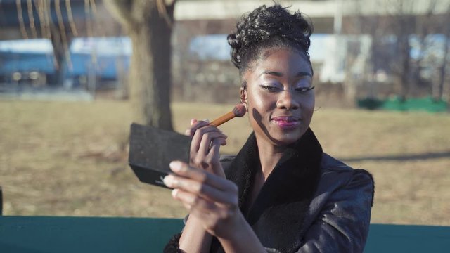 Woman applying make-up and blush while sitting on a bench in a park.