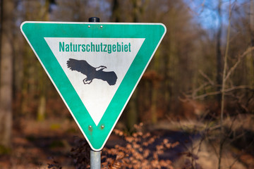 A triangular green and white sign on the edge of a forest. The sign indicates a nature reserve. This area is particularly sensitive and must be protected. Trees in the background.