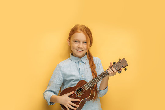 beautiful caucasian girl learns to play ukulele isolated over yellow background. attractive child with natural red hair hold small guitar, ukulele in hands and look at camera, smile, enjoy music