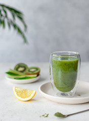 colorful green smoothie with spinach, celery, kiwi, lemon and green superfood powder in a double wall glass. vegan food. gray background with green leaves. vertical image. selective focus.