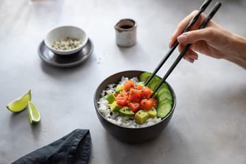 woman hand eating chopsticks hawaiian poke bowl with salmon, rice, avocado and cucumber. Diet food. horizontal image, gray background.