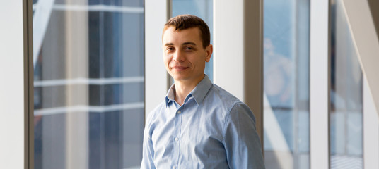 A 35-40 year old man in blue shirt poses in the office back hallway against the background of a large window. Portrait of a positive man near large office windows.