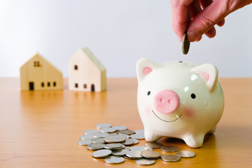 Selective focus of Men hand puting coin on piggy bank for saving for home, property with small wood house on the background. Wealth and saving plan for house mortgage. Personal investment concept.