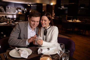 attractive caucasian lady show something interesting on smartphone to boyfriend. guy look at screen of mobile phone and smile, laugh. happy couple in restaurant