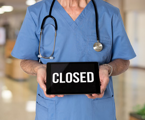 Senior male caucasian doctor with stethoscope in medical scrubs holding electronic tablet for saying hospital is closed due to coronavirus - Powered by Adobe