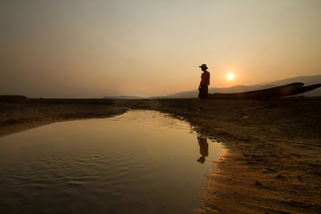 Young man standing near a drying river with a wooden boat at the place that once had been a big...