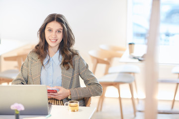 Candid portrait of pretty young woman looking at camera and holding smartphone while working or studying sitting at table in empty cafe, copy space