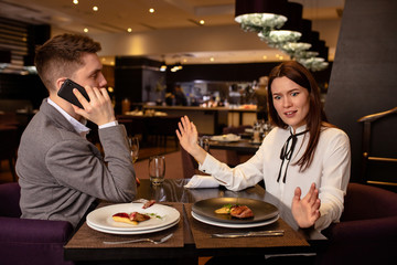 dissatisfied offended caucasian woman and handsome guy in restaurant. couple have quarrel, offended man and woman sit together, discuss relationships problems. man talk on phone