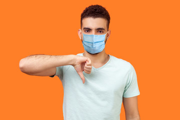 Thumbs down! Portrait of frustrated dissatisfied man with medical mask in casual white t-shirt showing dislike gesture, disapproval negative sign. indoor studio shot isolated on orange background