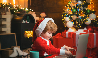 On-line Christmas shopping for kids. Happy little smiling boy wearing Santa clothes and writing a letter on his laptop. Fireplace background. Holidays. Little kid is wearing Santa clothes.