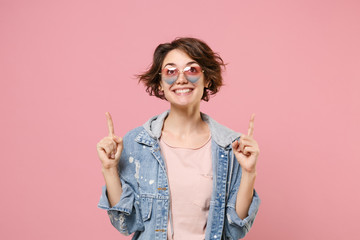 Cheerful young brunette woman girl in casual denim jacket eyeglasses posing isolated on pastel pink wall background in studio. People lifestyle concept. Mock up copy space. Pointing index fingers up.