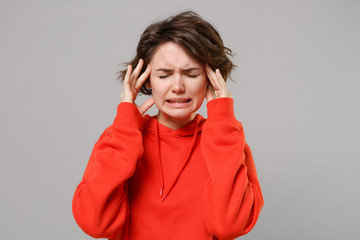 Sick young brunette woman girl in casual red hoodie posing isolated on grey background in studio. People lifestyle concept. Mock up copy space. Put hand on head, keeping eyes closed, having headache.