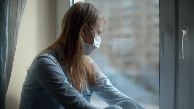 Young beautiful woman in home clothes and a protective mask sitting on the windowsill and looking out the window. Quarantine during the coronavirus epidemic. COVID-19 pandemic