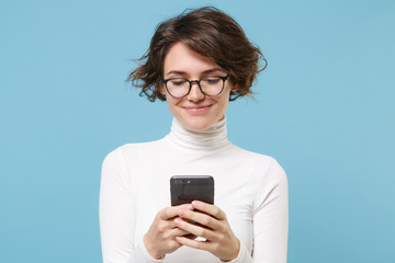 Fototapeta premium Smiling young brunette woman girl in casual white clothes, eyeglasses posing isolated on pastel blue background. People lifestyle concept. Mock up copy space. Using mobile phone, typing sms message.