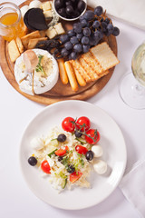 vegetables salad on the plate, glass of white wine and plate with assorted cheese, fruit and other snacks.
