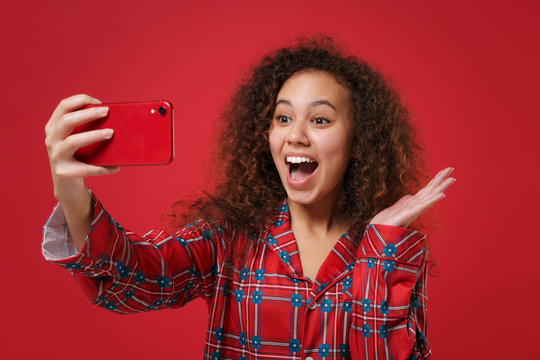 Excited young african american girl in pajamas homewear posing resting at home isolated on red background. Relax good mood lifestyle concept. Mock up copy space. Doing selfie shot on mobile phone.