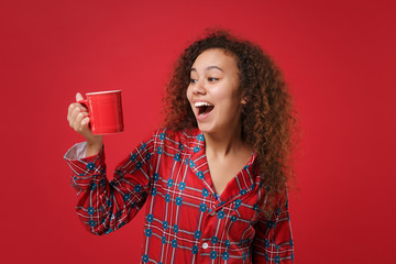 Surprised young african american girl in pajamas homewear posing while resting at home isolated on red background. Relax good mood lifestyle concept. Mock up copy space. Holding cup of coffee or tea.