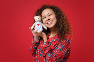Pretty young african american girl in pajamas homewear posing while resting at home isolated on red wall background. Relax good mood lifestyle concept. Mock up copy space. Hold teddy bear plush toy.