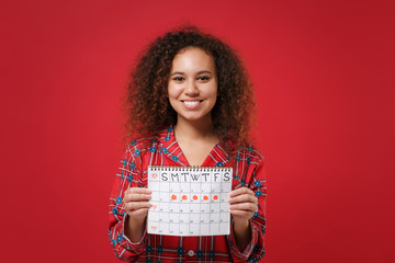 Smiling young african american girl in pajamas homewear posing rest at home isolated on red background. Relax good mood lifestyle concept. Hold female periods calendar for checking menstruation days.