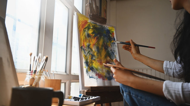 Closeup young beautiful artist girl concentrated on painting while sitting in front a drawing canvas over modern and orderly workshop as background.