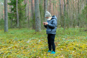 Kid with mask in their mouths due to coronavirus disease and play mobile games on a black phone in a forest
