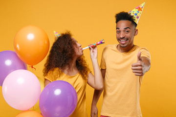 Smiling young friends couple african american guy girl in birthday hat isolated on yellow wall background. Holiday party concept. Celebrate hold colorful air balloons blowing in pipe showing thumb up.
