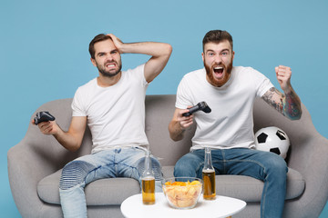 Displeased excited men guys friends in white t-shirt sit on couch isolated on blue background. Sport leisure concept. Cheer up support favorite team with soccer ball play game with joystick screaming.