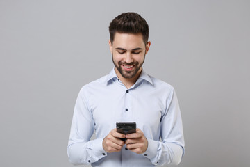 Smiling young unshaven business man in light shirt posing isolated on grey background in studio. Achievement career wealth business concept. Mock up copy space. Using mobile phone, typing sms message.