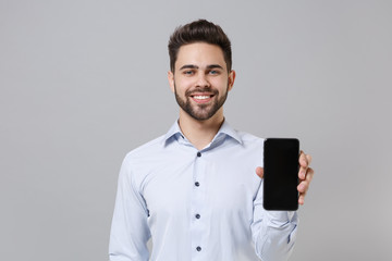 Smiling young unshaven business man in light shirt posing isolated on grey wall background. Achievement career wealth business concept. Mock up copy space. Hold mobile phone with blank empty screen.