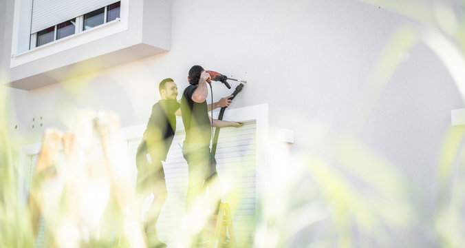 zwei freundliche Handwerker bohren mit der Bohrmaschine in die Hauswand um sp&auml;ter eine Markise zu befestigen