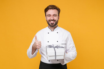 Smiling young bearded male chef cook or baker man in white uniform isolated on yellow background. Cooking food concept. Mock up copy space. Hold present box with gift ribbon bow, showing thumb up.