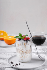 Overnight oats with yogurt topped with berries in a glass jar. Coffee and oranges in the background