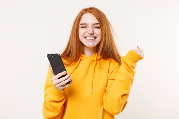 Happy young redhead woman girl in yellow hoodie posing isolated on white background studio portrait. People emotions lifestyle concept. Mock up copy space. Using mobile phone, doing winner gesture.