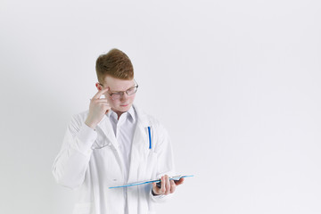 Young trainee doctor intern stands on white background. Handsome red- haired man, thinking