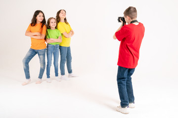 Fototapeta premium A boy photographer takes pictures of three cute girls in bright T-shirts on a white background in the studio. Young photographer and blogger.