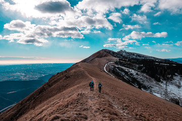 Walking to heaven. In a autumnal landscape, a meadow with dry (brown) grass, a blue sky with clouds and trekkers along the path and the mountain ridge. Col Visentin, Nevegal, Italian dolomites, Italy