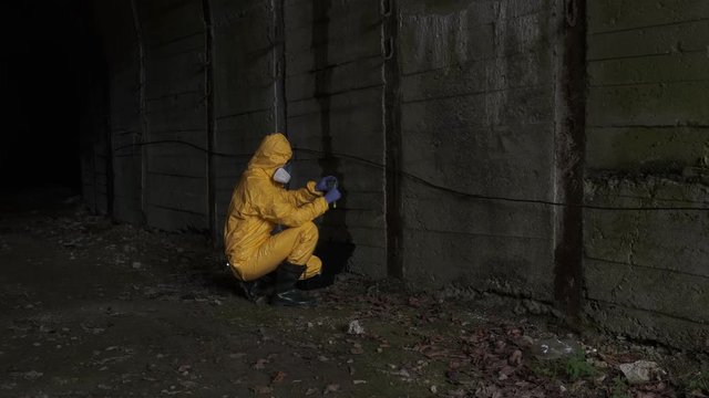 A man in a yellow protective suit with a research bag picks up soil samples for research in a dark tunnel.