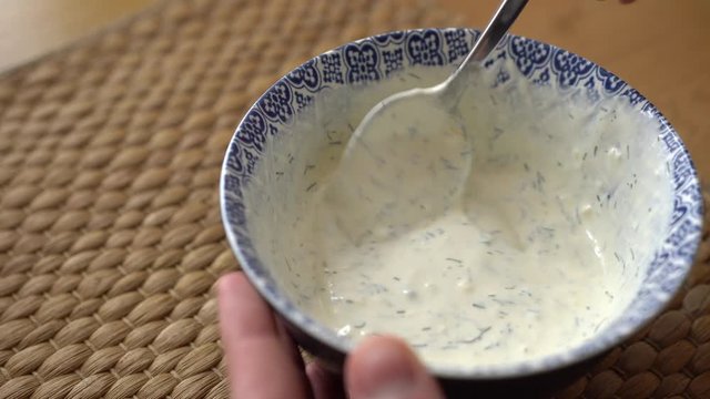Mixing garlic sauce with a tablespoon. Garlic dip in a bright white and blue bowl on the wooden table. Closeup of a small pot of garlic mayonnaise mixing with a spoon.