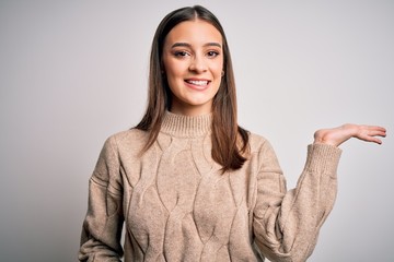 Obraz premium Young beautiful brunette woman wearing casual sweater standing over white background smiling cheerful presenting and pointing with palm of hand looking at the camera.