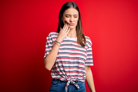 Young beautiful brunette woman wearing casual striped t-shirt standing over red background touching mouth with hand with painful expression because of toothache or dental illness on teeth. Dentist