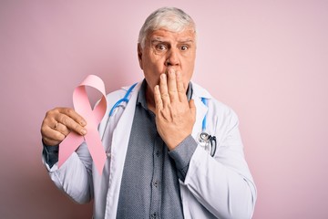 Senior handsome hoary doctor man wearing stethoscope holding pink cancer ribbon cover mouth with hand shocked with shame for mistake, expression of fear, scared in silence, secret concept