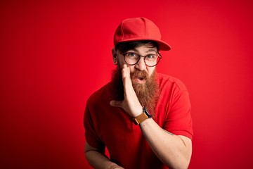 Young handsome delivery man wearing glasses and red cap over isolated background hand on mouth telling secret rumor, whispering malicious talk conversation